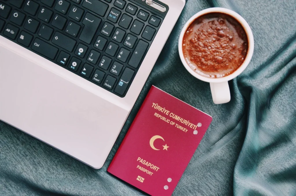 A passport is sitting next to a laptop and a cup of coffee.